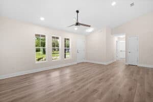 An empty 4 bedroom room with light wood-look flooring, pale pink walls, a ceiling fan, and three windows overlooking a yard. Townsley Construction in Florida