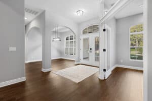 A foyer in this 3-bedroom house features wood floors, a light-colored area rug, a glass-paned double front door, and an arched entryway leading to a dining area. Townsley Construction in Florida