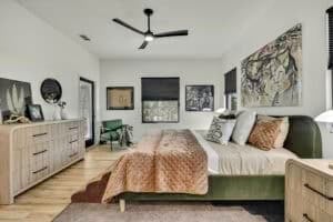 A Harper bedroom featuring a green bed with a tan quilted coverlet, two light-wood nightstands, a ceiling fan, a green armchair, and several pieces of wall art. Townsley Construction in Florida