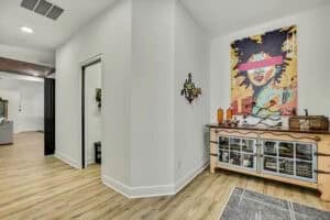 A hallway with light wood floors in The Harper features a buffet cabinet topped with a large, colorful portrait painting of a woman with a pink bar across her eyes. Townsley Construction in Florida