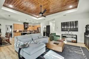 Open-concept living room and kitchen in The Harper featuring a light gray sectional sofa, a wood coffee table, and a wooden tray ceiling with a black ceiling fan. Townsley Construction in Florida