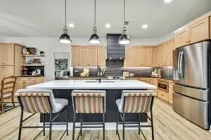 Modern kitchen in The Harper featuring light wood cabinets, a black hood vent, stainless steel appliances, and a central island with three upholstered stools. Townsley Construction in Florida