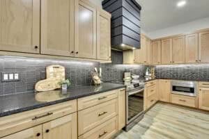 A kitchen in The Harper featuring light wood cabinets, dark granite countertops, a dark tile backsplash, and stainless steel appliances. Townsley Construction in Florida