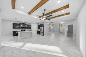 Open-concept kitchen and living area in the Bobak floor plan featuring dark cabinets, a white kitchen island, wood ceiling beams, and light-colored tile floors. Townsley Construction in Florida