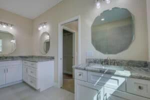 A bathroom in The Harper with white double vanities, marble countertops, and two octagonal mirrors, featuring an open doorway in the center that reveals a walk-in closet. Townsley Construction in Florida