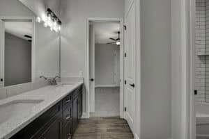 A bathroom vanity with a white speckled countertop and dark cabinets in The Purvis, showing a view through a doorway into a bedroom and a glimpse of a tiled shower area. Townsley Construction in Florida