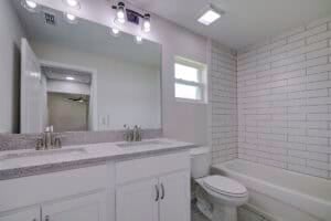 Bathroom in a Houston 4-2-2 floor plan featuring a double vanity with granite-style countertops, a large mirror, a toilet, and a bathtub with white subway tile surround. Townsley Construction in Florida