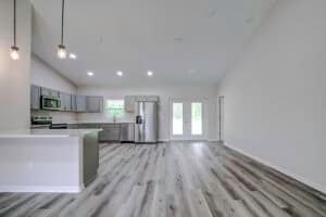 An open-concept kitchen and living area in this 4 bedroom house features gray cabinetry, stainless steel appliances, and light wood-look flooring leading to a pair of glass patio doors. Townsley Construction in Florida