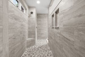 A walk-in shower in this 3 bedroom house features light grey tiled walls, a small built-in bench, two wall niches, and a mosaic tiled floor. Townsley Construction in Florida