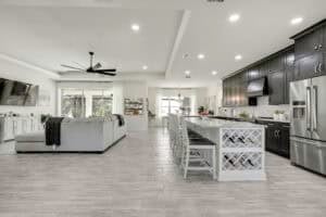 Open-concept kitchen and living area for this new home floor plan, featuring a large grey sectional sofa, a white kitchen island with bar seating and built-in wine storage, and black cabinetry. Townsley Construction in Florida