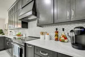 A kitchen with dark gray cabinets, white marble countertops, a white patterned tile backsplash, and a black coffee maker, part of a new home floor plan. Townsley Construction in Florida