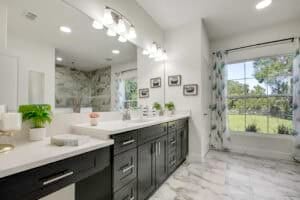 Bathroom with a double vanity featuring dark cabinets, a white countertop, and a large mirror reflecting a walk-in shower in this new home floor plan. Townsley Construction in Florida