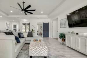 Living room in a new construction home with a large white sectional sofa, a patterned ottoman, and a television mounted above a console cabinet, featuring a blue sliding barn door in the background of this flex space floor plan. Townsley Construction in Florida