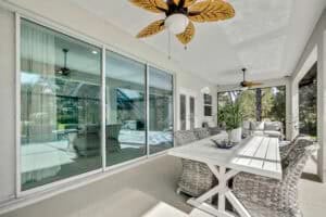 A covered patio in this 3 bedroom house features a white dining table with wicker chairs, a leafy plant centerpiece, and two ceiling fans with palm-leaf blades. Townsley Construction in Florida