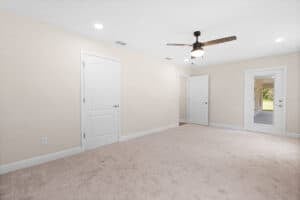 An empty room in this Houston home features beige walls, tan carpeting, a ceiling fan, two interior doors, and one door with a glass pane leading to a porch. Townsley Construction in Florida