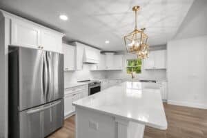A kitchen in the Hamblin floor plan featuring white cabinets, a stainless steel refrigerator, a central island with a white countertop, and a gold geometric chandelier. Townsley Construction in Florida