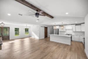 Open-concept kitchen and living area in the Hamblin floor plan, featuring white cabinets, a central island, a wood-beam ceiling, and wood-look flooring. Townsley Construction in Florida