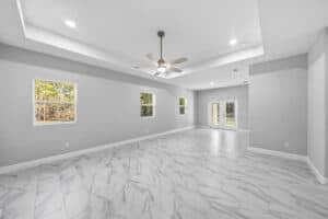 An empty, grey-walled flex space with a tray ceiling, ceiling fan, and white marbled tile flooring featuring windows and double glass doors in this Bobak home. Townsley Construction in Florida