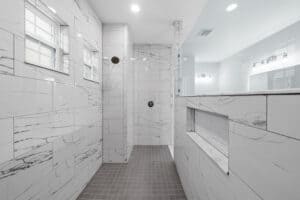 Walk-in shower in this 3 bedroom house featuring white marble-patterned wall tiles, a grey square-tiled floor, and a built-in recessed niche. Townsley Construction in Florida