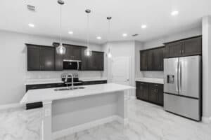 A modern kitchen in this 3 bedroom home features dark wood cabinets, a white marble-topped island with a sink, stainless steel appliances, and marble-look tile flooring. Townsley Construction in Florida
