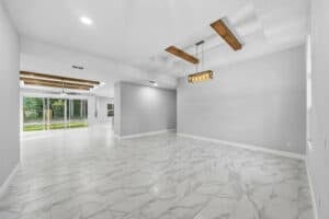 An open-concept living area in this 3-bedroom Bobak home featuring white marble-look tile flooring, gray walls, and exposed wooden ceiling beams. Townsley Construction in Florida