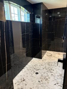 A walk-in shower in the Wyrick floor plan featuring black-and-gold veined tile walls, a light-colored mosaic-tiled floor, an arched window, and a shower head. Townsley Construction in Florida