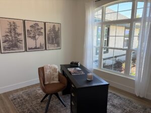 A home office in The Wyrick featuring a black desk and brown swivel chair, with three botanical prints on the wall and a large window overlooking a yard. Townsley Construction in Florida