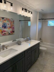 Double vanity bathroom in this Wyrick floor plan home with dark gray cabinets, a white stone countertop, dual sinks, and a large mirror beneath two four-light fixtures. Townsley Construction in Florida