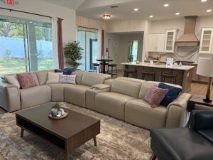 A large tan leather sectional sofa faces a wooden coffee table in the open-concept living area of this Wyrick floor plan home, with a kitchen visible in the background. Townsley Construction in Florida
