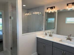 A modern bathroom in The Bailey featuring a double vanity with grey cabinets, a quartz countertop, and a glass-enclosed shower. Townsley Construction in Florida
