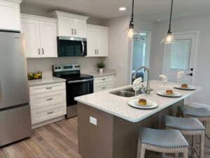 A modern kitchen in The Bailey, featuring white cabinets, stainless steel appliances, and a central island with gray bar stools and place settings. Townsley Construction in Florida