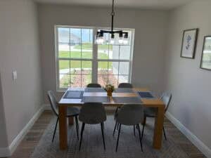 A wooden dining table with six gray chairs sits on a patterned rug in front of a window in The Bailey. Townsley Construction in Florida