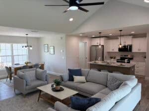 An open-concept living area in The Bailey featuring a grey sectional, a coffee table with a small plant, and an adjacent kitchen with a breakfast island and dining table. Townsley Construction in Florida