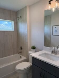 A dark vanity, white countertop, toilet, and tiled shower-tub combination in one of the bathrooms of The Bailey 2 bath home. Townsley Construction in Florida