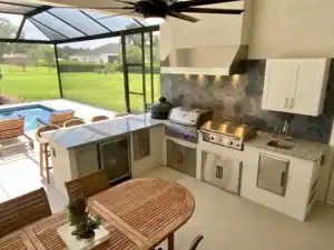 Outdoor kitchen on a screened-in patio of this 4 bedroom house, featuring a built-in grill, smoker, stainless steel cabinets, and a granite countertop, overlooking a backyard swimming pool. Townsley Construction in Florida