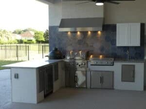Outdoor kitchen in The Adkin featuring a grill, a ceramic smoker, a flat-top griddle, and a sink, set against a blue hexagonal tile backsplash. Townsley Construction in Florida