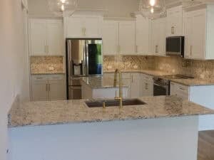 A kitchen in The Adkin featuring white cabinets, a central island with a gold faucet, stainless steel appliances, and marbled stone countertops. Townsley Construction in Florida
