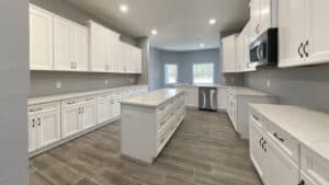 A modern kitchen in this 3-bedroom home features white cabinets, a central island, light-colored countertops, and grey wood-look tile flooring. Townsley Construction in Florida