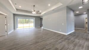 An open-concept living area in this 3 bedroom home features gray walls, wood-look tile flooring, a tray ceiling with a fan, and a large sliding glass door looking out to trees. Townsley Construction in Florida